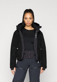 Černá bunda s texturovaným kordovým vnějším povrchem, huňatou podšívkou kapuce a předním zipem. Nosená přes černý top a tmavé kalhoty s kapsami.