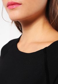 Maglione nero con scollo a barca, tessuto strutturato a tinta unita e cuciture laterali in velluto a contrasto. Dettaglio ravvicinato del colletto e del tono della pelle.