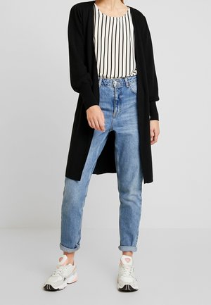 Langer schwarzer Cardigan über einem gestreiften weißen und schwarzen Top, kombiniert mit hellblauen, locker sitzenden Jeans und weißen Sneakers mit pinken Akzenten.