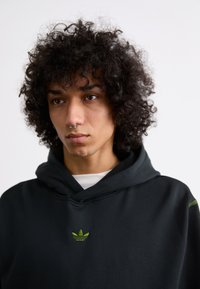 Svart hoodie med grön broderad logotyp på bröstet, mjukt material, med dragsko på huvan och ribbade manschetter, kroppsnära design.