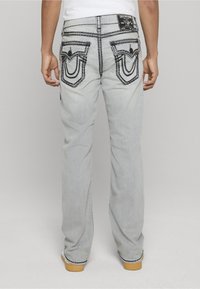 Vaqueros de color gris claro con costuras negras en contraste, que cuentan con dos bolsillos traseros con solapas decorativas y una parche con el logo en la cintura.