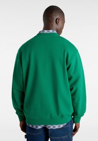 Sudadera verde de corte relajado, con mangas acampanadas y cuello redondo, superpuesta a una camisa de cuadros con patrones en azul y blanco.