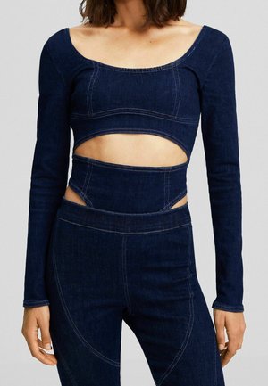 Kvinde iført en mørkeblå denim crop top med lange ærmer og matchende højtaljede bukser med udskæringer omkring taljen.
