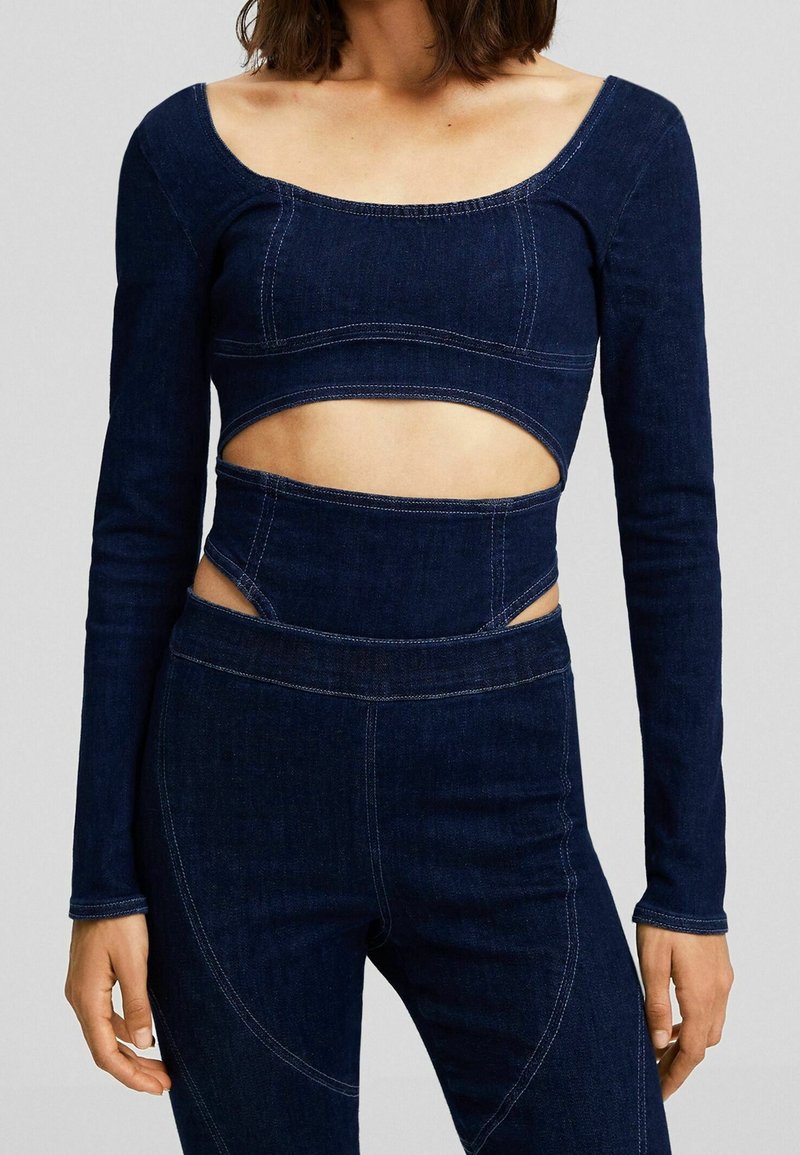 Femme portant un crop top en denim bleu foncé à manches longues et un pantalon taille haute assorti avec des découpes autour de la taille.