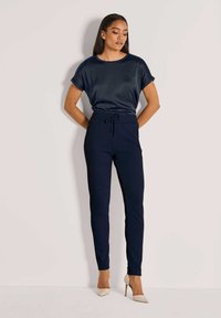 Vrouw in een marineblauwe blouse met korte mouwen en bijpassende hoog-geknoopte broek met beige hoge hakken, staand tegen een eenvoudige lichte muur.