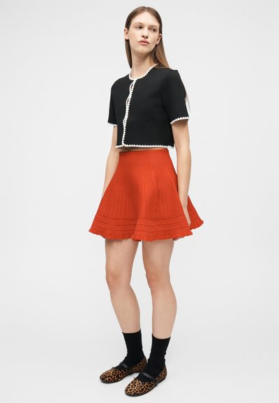 Γυναίκα που φοράει μαύρο κοντό μπλουζάκι με λευκή επένδυση, κόκκινη φουσκωτή φούστα, μαύρες κάλτσες και παπούτσια φλατ με λεοπάρ σχέδιο, στέκεται πλευρικά.