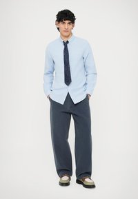 Světle modrá košile na knoflíky, tmavě modrá kravata se vzorem, tmavě modré kalhoty širokých nohavic a dvoubarevné boty. Pohodlný střih s hladkou látkou a viditelným šitím.