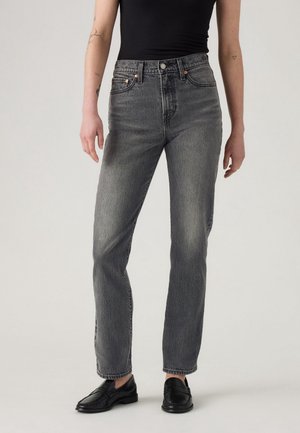 Personne portant un jean gris délavé taille haute, un débardeur côtelé noir, et des mocassins en cuir noirs, debout devant un fond clair uni.