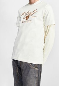 Valkoinen puuvillatee-paita, jossa on ruskea graafinen printti, jossa lukee "Flight" ja "Jordan", kerroksittain vaaleansinisen pitkähihaisen paidan päällä.