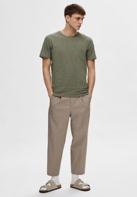 T-shirt olive vert à manches courtes, pantalon beige ajusté, chaussettes blanches à la cheville et sandales grises claires à deux brides, se tenant sur un fond neutre.
