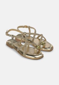 Sandalias planas doradas con tiras entrecruzadas de material brillante, lazo para el dedo del pie y acentos decorativos de hardware. Suela plana y suave.