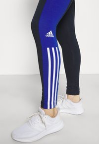 Legginsy Adidas w kolorze niebieskim i czarnym z białymi paskami akcentującymi oraz logo; w połączeniu z białymi butami sportowymi z teksturowanym materiałem.