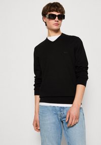 Schwarzer Strickpullover mit V-Ausschnitt und gerippten Bündchen, kombiniert mit hellblauen Jeans. Schwarze rechteckige Sonnenbrillen ergänzen das Outfit.