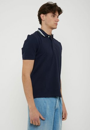 Jonge man in een marineblauw poloshirt met gestreepte kraag en lichtblauwe spijkerbroek, staand in profiel tegen een effen witte achtergrond.