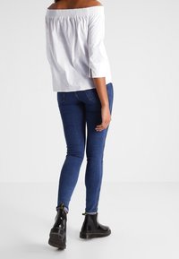 Top bianco a spalle scoperte con maniche a tre quarti, abbinato a jeans skinny blu scuro e stivaletti neri con dettagli gialli.