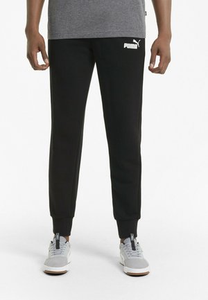 Mężczyzna w czarnych dresach Puma z białym logo na udzie, szarych butach sportowych i szarej koszulce, stojący na białym tle.