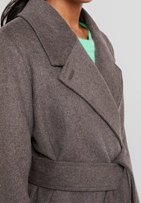 Grau melierter Wollmantel mit weit geschnittenem Kragen, tailliertem Bund und strukturiertem Finish. Sichtbare umgeschlagene Revers über einem hellgrünen Pullover darunter.