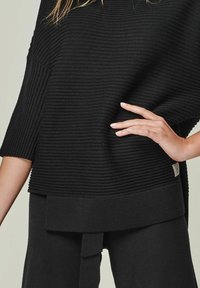 Schwarzer gerippter Strickpullover mit Dreiviertelärmeln, der einen breiten Saum und ein kleines Etikett-Detail an der Seite aufweist. Nahaufnahme der Stoffstruktur.