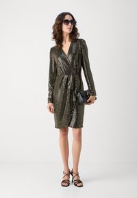 Robe en sequins noire et dorée avec des manches longues et un décolleté en V, présentant un design drapé et une taille cintrée. Associée à des talons noirs à lanières.