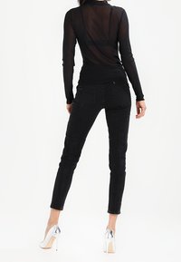 Žena v průhledné černé blůze s dlouhými rukávy, černých skinny džínách a stříbrných lodičkách, stojící zády k fotoaparátu na bílem pozadí.