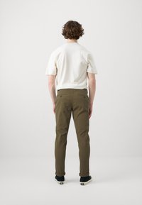 Beige kortärmad skjorta, olivgröna slim-fit byxor, rullade cuffs, stylad med mörka sneakers, sedd bakifrån mot en neutral bakgrund.