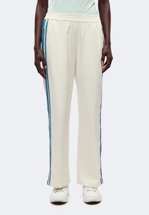 Personne portant un pantalon de survêtement couleur crème avec des bandes latérales bleues et des baskets blanches, debout devant un fond clair uni.