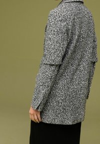 Abrigo texturizado en gris y negro con un cuello entallado y bolsillos laterales, que presenta un corte holgado y mangas hasta los codos.