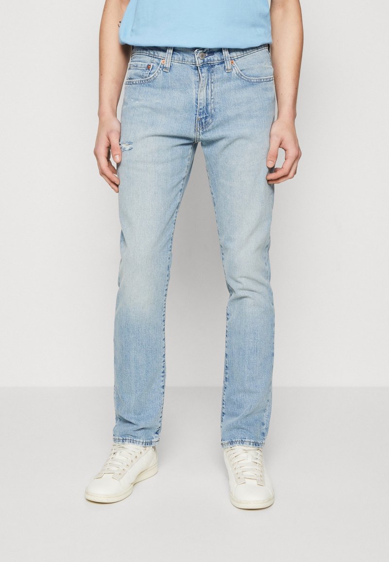 Jeans en denim bleu clair avec une coupe slim, présentant un léger effet usé, cinq poches et une fermeture classique à boutons. Portés avec des baskets blanches.