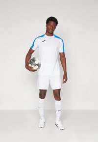 Mees valges jalgpallikomplektis, millel on sinised triibud, hoiab must-valget jalgpallivoodi ja seisab ühtlase valge tausta ees.