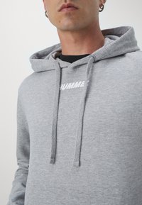 Uomo che indossa una felpa con cappuccio grigio chiaro con la scritta "HUMMEL" parzialmente visibile sul petto, indossa anche orecchini a cerchio in argento e una maglietta nera sotto.