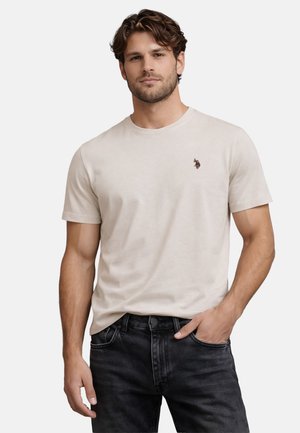 Man with short brown hair wearing a beige short-sleeve t-shirt with a small logo on the chest and black jeans, hand in pocket.