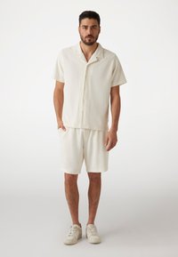Hellbeige kurzärmliges Hemd mit Kragen, kombiniert mit passenden Shorts, beide aus einem strukturierten, gerippten Stoff. Getragen mit weißen Sneakers.