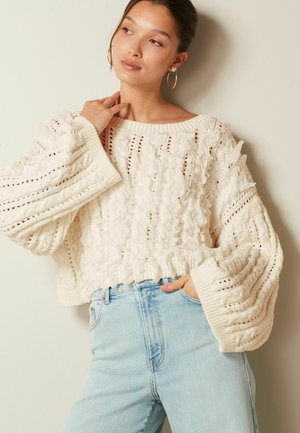 Kreemikaalune kudumissweater volangdetailidega, soonikkoes mansetid ja avatud mustriga, koos helesiniste kõrge vöökohaga teksadega.