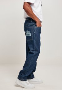Blue denim jeans with a relaxed fit, featuring a large light blue rainbow graphic on the back pocket. Worn with white sneakers.
