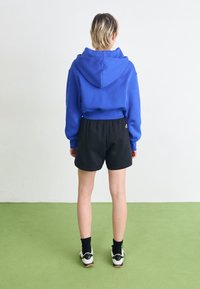 Sudadera con capucha azul, pantalones cortos negros y zapatillas blancas con detalles en negro. La modelo mira hacia otro lado, mostrando el ajuste casual y los materiales.