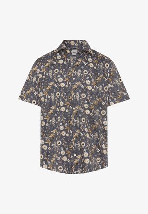 Mann mit leichtem Gesichtshaar, der ein dunkelbraunes Poloshirt unter einem blau geblümten Hemd vor weißem Hintergrund trägt.