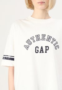 Bílý bavlněný tričko s nápisem "AUTHENTIC GAP" v tučných černých písmenech, s námořnickými pruhy na rukávech a volným střihem.