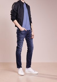 Svart bomberjacka, blå t-shirt, mörka jeans och vita sneakers. Outfitet har en avslappnad passform och släta texturer.