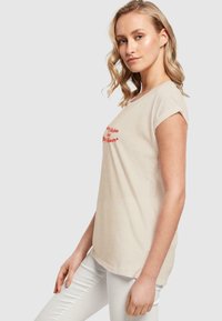 Beige Baumwoll-T-Shirt mit umgeschlagenen Ärmeln und rotem Text "Bleib immer ambitioniert und glücklich." Lockerer Schnitt, leichtes Material, lässiger Stil.