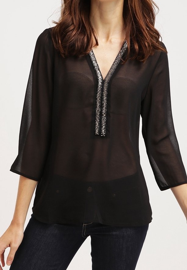 Donna che indossa una blusa nera trasparente con maniche 3/4 e rifiniture in perline argentate lungo il collo a V, abbinata a jeans scuri.