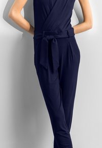 Námorní jumpsuit s překládaným designem, samostatně vázaným opaskem v pase a plisovanými detaily v přední části. Vyroben z měkké, lehké tkaniny s hladkou strukturou.