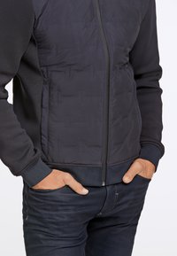 Svart atletisk jacka med quiltad framsida, ribbat manschett och nederkant. Har dragkedjestängning och två sidofickor. Bärs över mörka jeans.