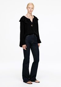 Frau mit blonden Haaren, die eine schwarze Bluse mit gerafftem Kragen, dunkelblaue Jeans, schwarze Sandalen und eine runde Anhängerkette trägt.