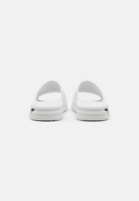 Scarpe bianche slip-on con tallone aperto, texture liscia, design leggermente curvato e suola spessa con un piccolo dettaglio a intaglio.