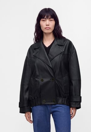 Frau mit langen dunklen Haaren, die eine schwarze, zweireihige Lederjacke und blaue Jeans trägt, steht vor einem schlichten hellen Hintergrund.