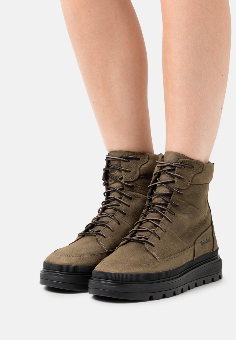 Bottes en cuir vert olive à tige haute, finition texturée, embout renforcé et semelles en caoutchouc noir. Elles comportent des lacets et des œillets marron.