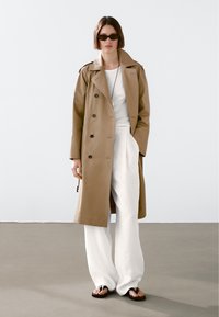 Beżowy trencz o podwójnym zapięciu, czarne guziki, w zestawieniu z białymi spodniami o szerokich nogawkach i czarnymi klapkami. Minimalistyczny styl.