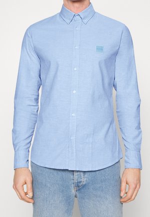 Mann trägt ein hellblaues Hemd mit Knöpfen und einem kleinen quadratischen "BOSS"-Logo auf der linken Brust, kombiniert mit hellblauen Jeans.