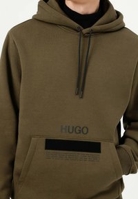 Oliwkowa bluza z kapturem wykonana z miękkiego materiału, z czarnym sznurkiem, kieszenią z przodu oraz nadrukowanym logo i tekstem z przodu.