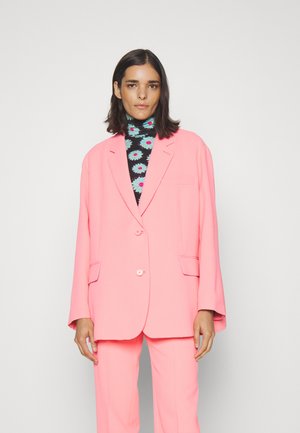 Per didelį rožinį švarką su dviem mygtukais, priekinių kišenių ir smailiais atlapais, dėvimas juodas megztinis su spalvingu gėlių raštu.